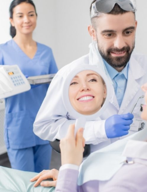 young-smiling-woman-looking-mirror-after-procedure-professional-whitening-while-sitting-armchair-dentist-office